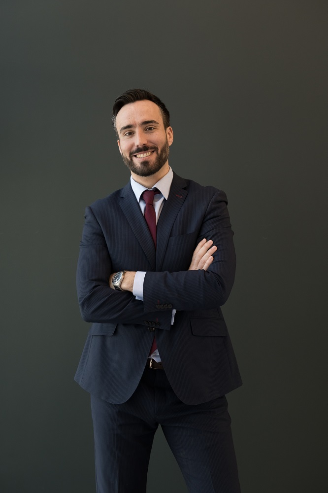 Smiling unshaven male in black business attire with wristwatch looking at camera standing with folded arms over black background