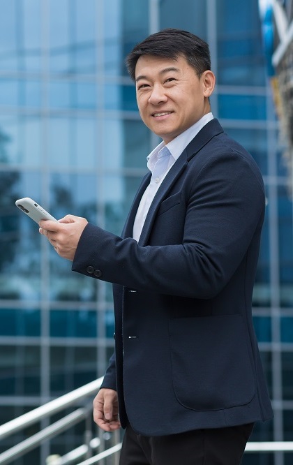 Portrait of an Asian businessman outside an office building, a man in a business suit is using a smartphone, smiling and looking at the camera, walking down the street.