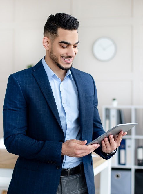 Portrait of young Arab man in formal suit working online, using digital tablet in modern office. Millennial Eastern guy reading documents, checking email or browsing web for information on touch pad